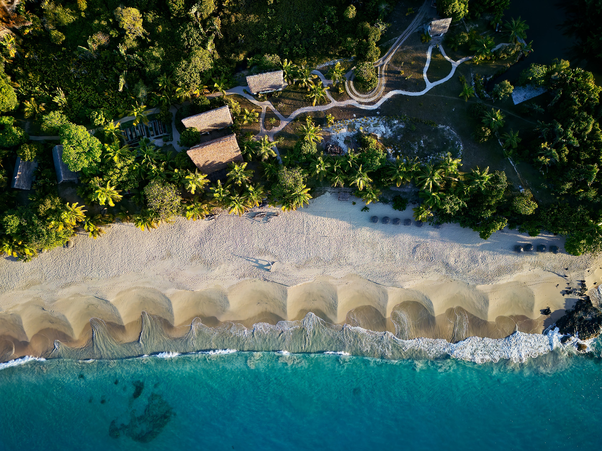 Aerial view of tropical beach with thatched-roof huts, palm trees, white sand, and turquoise ocean waves.