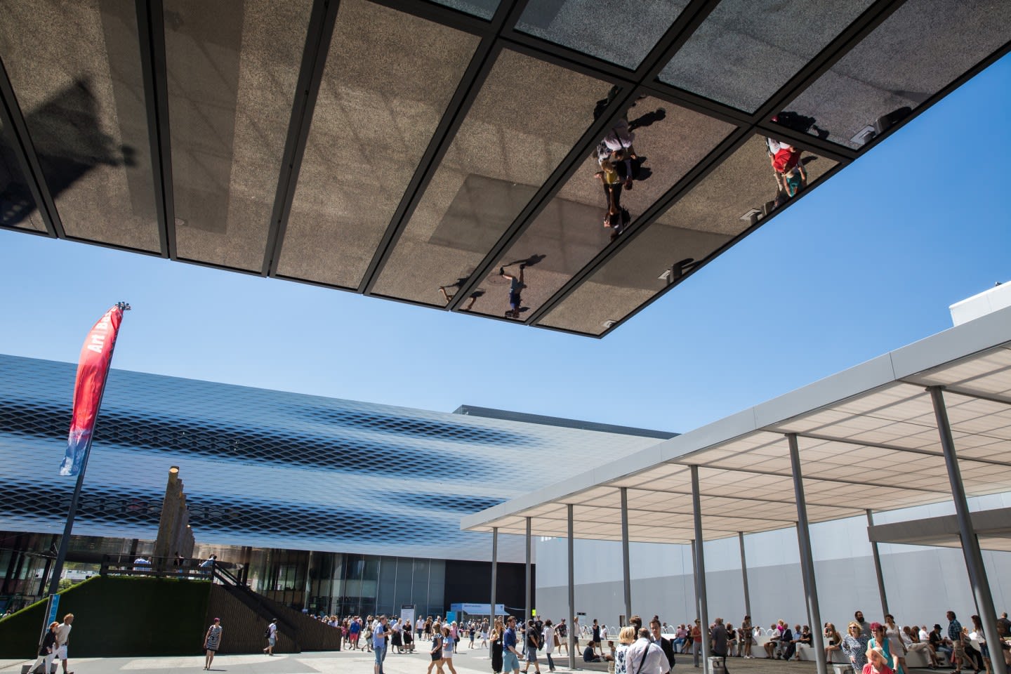 Crowds gather under reflective canopy at Art Basel, with red banner and modern architecture against blue sky