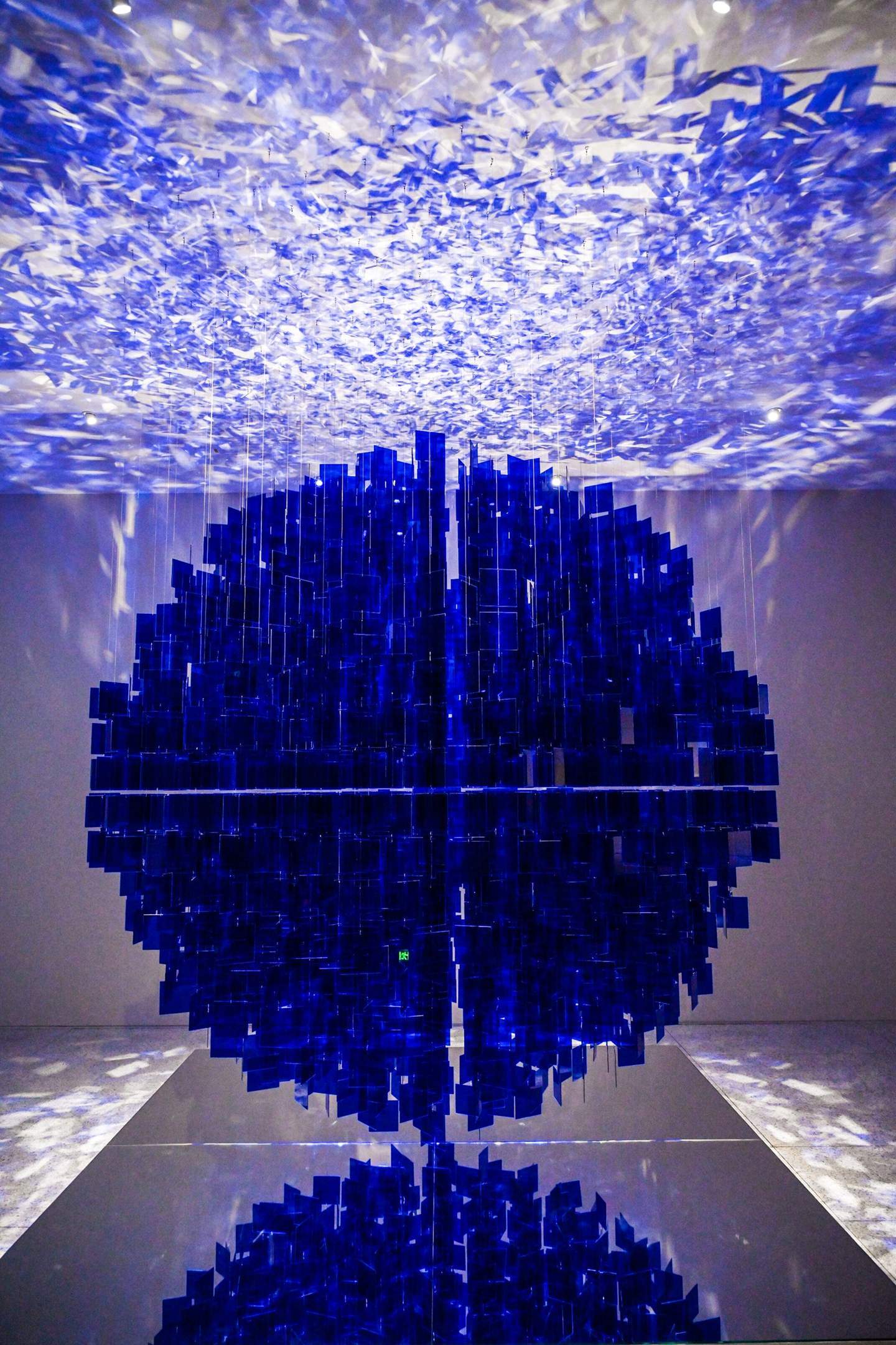 Suspended cobalt blue glass sphere installation reflected on mirrored floor with shimmering light patterns on ceiling