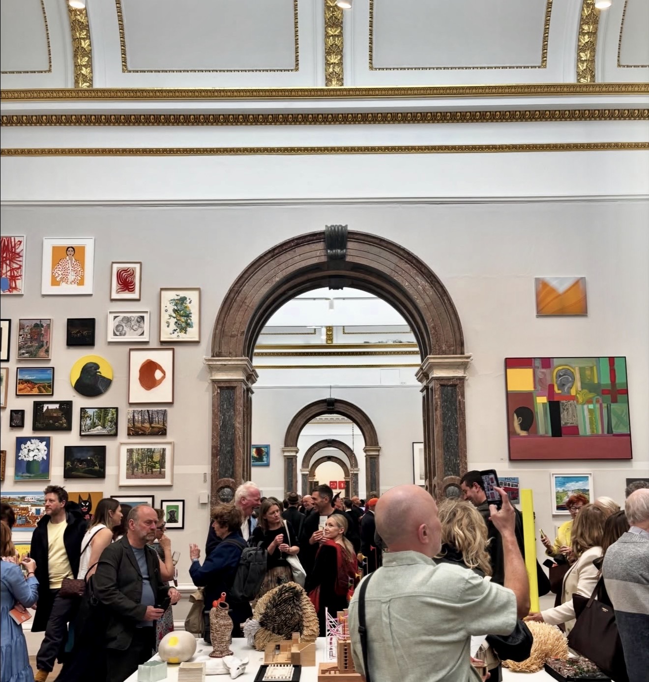 Crowd of visitors at a busy art exhibition with salon-style hung paintings and a grand marble archway