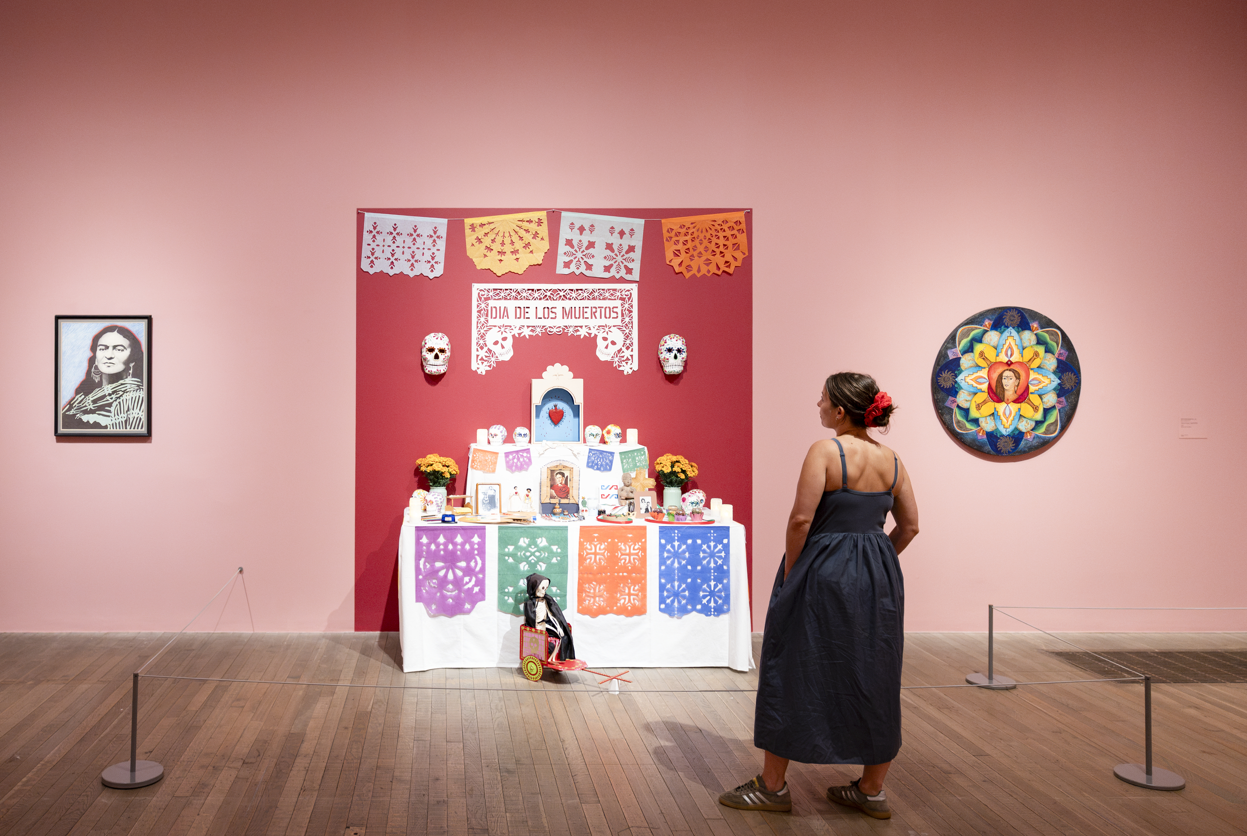 Woman in navy dress views Dia de los Muertos altar at Frida: The Making of an Icon exhibition, flanked by Frida Kahlo artworks