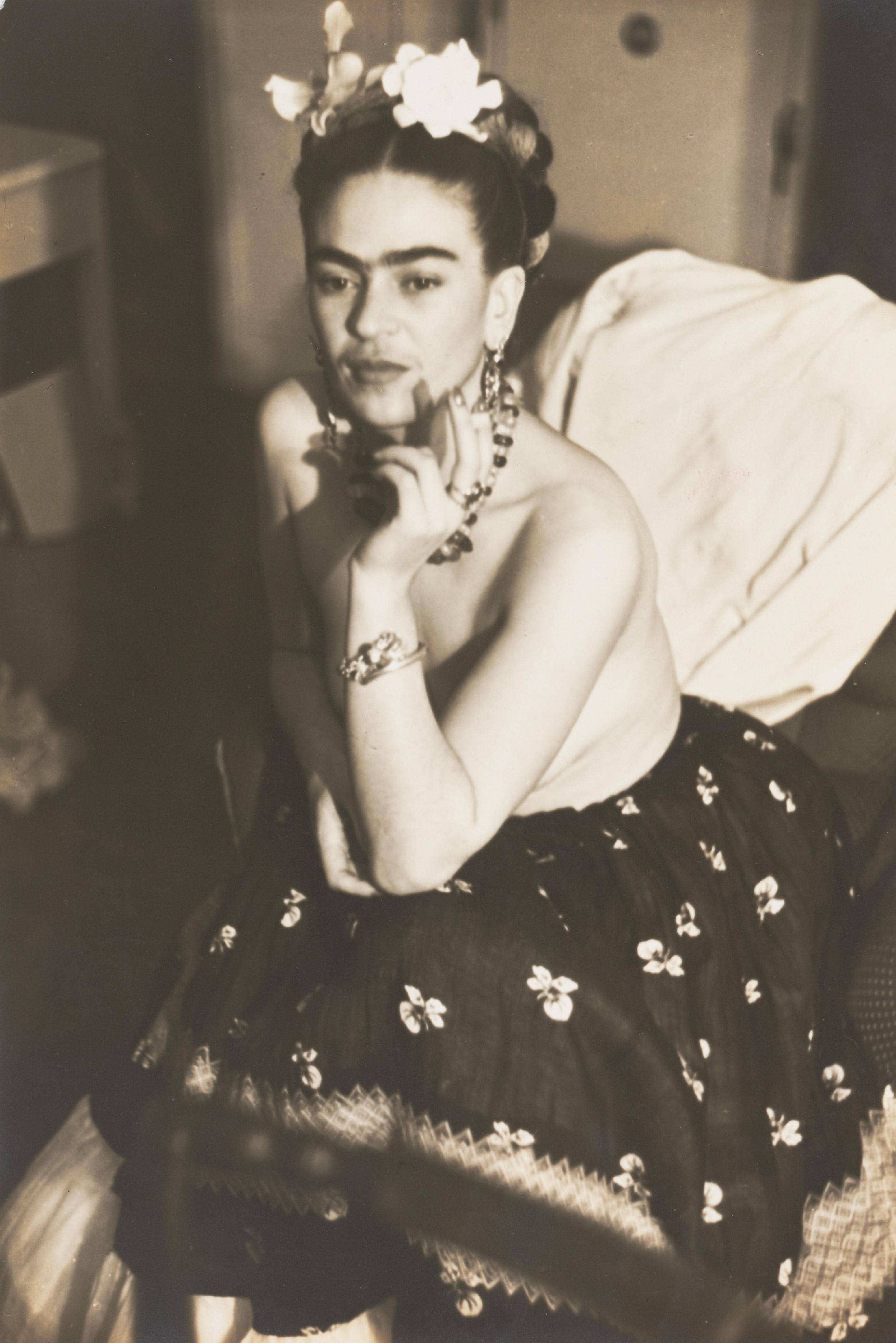Vintage sepia photograph of Frida Kahlo by Julien Levy, wearing flowers in her hair and a floral skirt, hand to chin