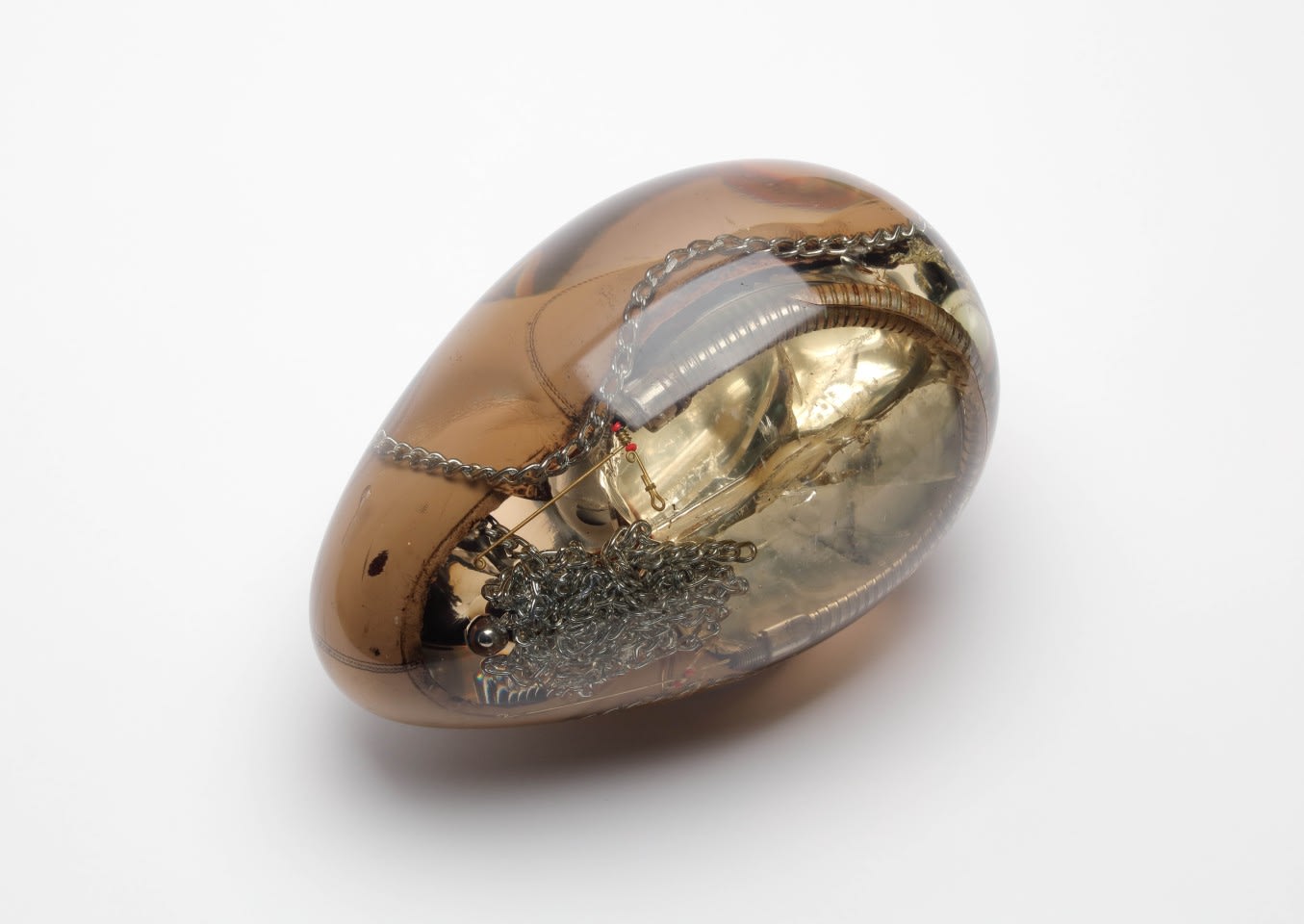 Egg-shaped translucent resin sculpture by Nakanishi encasing tangled metal chains, wires and beads, on white